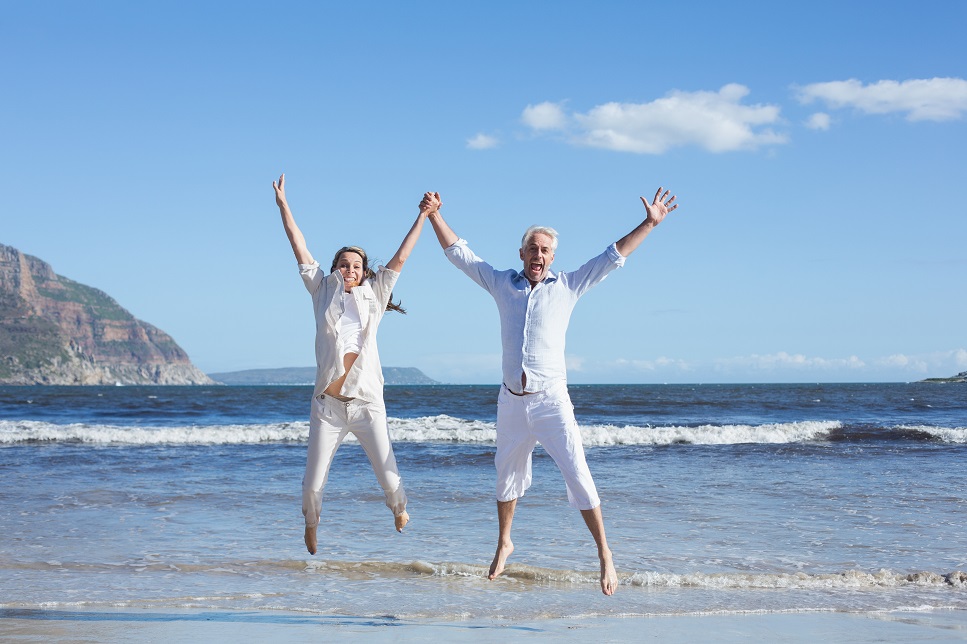 Retirement Travel - Happy couple jumping up barefoot on the beach on a sunny day